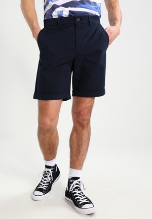 Marineblauwe katoenen korte broek met opgerolde zoom, steekzakken aan de voorkant en een knoopsluiting, gecombineerd met zwarte hoge sneakers en witte sokken.