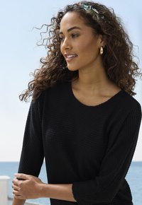 Schwarzer Waffelpullover mit Dreiviertelärmeln. Das Model hat lockiges Haar und trägt goldene Creolen sowie ein gemustertes Stirnband.