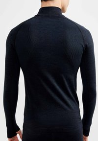 Donker langmouwtop met een hoge kraag, voorzien van textuurpaneel en gladde stof, ontworpen voor sportkleding. Naaddetails zichtbaar.