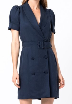 Vrouw in een marineblauwe blazerjurk met korte mouwen, een ceintuur, gewatteerde schouders en dubbele knoopdetails.