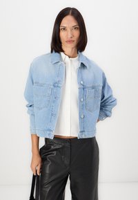 Giacca di jeans azzurro chiaro con design cropped, dotata di due tasche frontali, chiusura con bottoni e una vestibilità morbida. Indossata sopra una maglietta bianca.