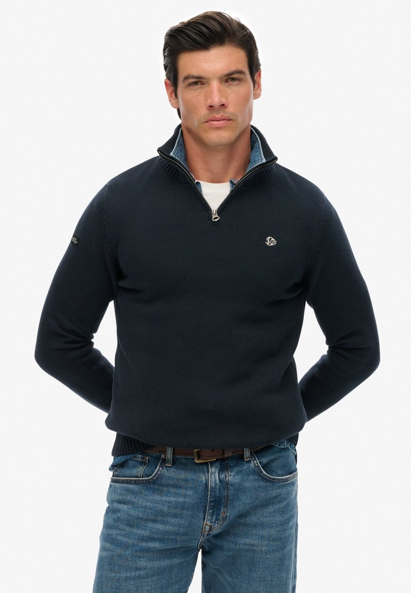 Marineblauer Strickpullover mit halbhohem Reißverschlusskragen, dezentem Logo und gerippten Bündchen, kombiniert mit hellblauen Jeans.