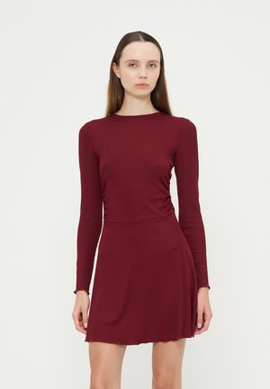 Jonge vrouw met lang bruin haar, gekleed in een strak vallende lange mouwen bordeauxrode jurk, staande tegen een effen witte achtergrond.