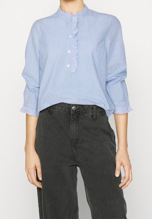 Modella che indossa una blusa a righe blu chiaro con colletto arricciato e jeans neri a vita alta, con le mani appoggiate sulla vita.
