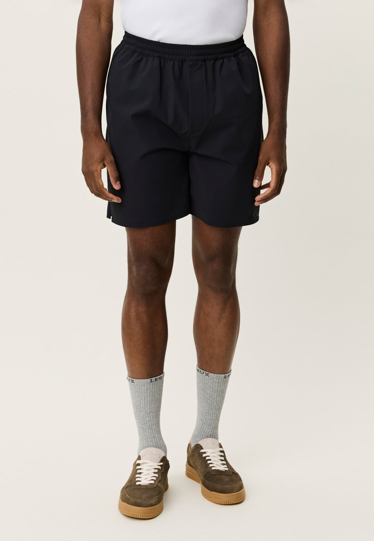 Shorts de sport noirs en matériau léger avec une ceinture élastique. Portés avec des chaussettes grises et des baskets marron.