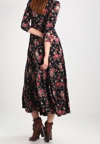 Donna che indossa un vestito midi nero a fiori con maniche a tre quarti e stivaletti con tacco marroni, in piedi con le mani dietro la schiena.