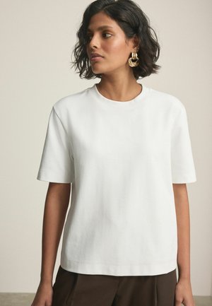 Kvinde med kort mørkt hår iført en simpel hvid t-shirt med korte ærmer og store gyldne hoop-øreringe, der kigger til venstre mod en neutral baggrund.