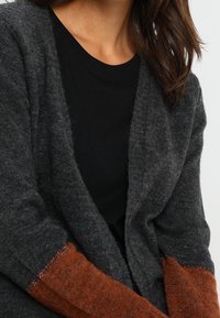 Grå cardigan med en mjuk textur, har långa ärmar i en orange-brun färg. Designen är öppen framtill med en avslappnad passform.