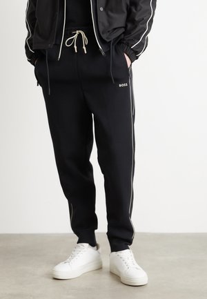 Pantalons de jogging noirs avec taille élastique et cordon de serrage blanc, présentant des rayures sur les côtés et un petit logo. Associés à des baskets blanches.