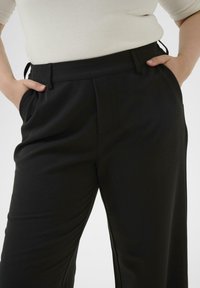 Črne krojene hlače z gladkim materialom, ravnim krojem nogavic in stranskimi žepi. Dodatki vključujejo čisto pasnico in skrite zapirala.