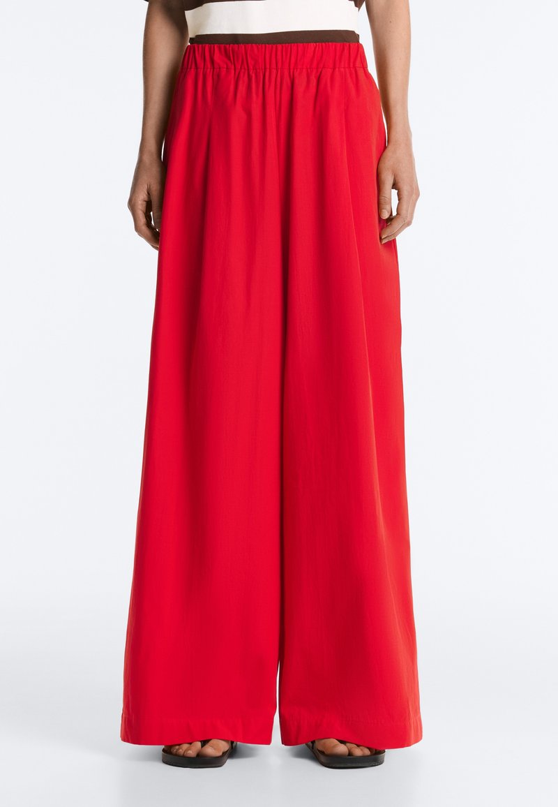 Personne portant un pantalon large rouge vif avec une taille élastique et des sandales noires sur un fond uni.