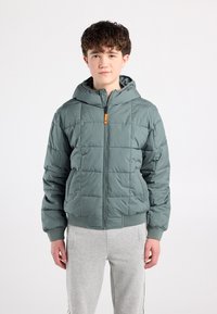 Gewatteerd groene jas met capuchon, voorzien van een ritssluiting en geribbelde manchetten. Het ontwerp heeft een doorgestikte textuur en een oranje accent op de rits.