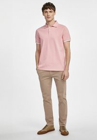 Różowa koszulka polo z białymi akcentami, zestawiona z beżowymi chinosami i brązowymi butami. Luźny krój, gładka tekstura, klasyczny design kołnierzyka.