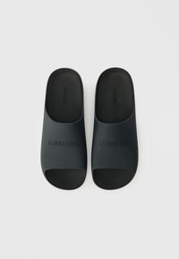 Μαύρες καουτσούκ σαγιονάρες με ανοιχτή μύτη, διαθέτοντας ανάγλυφο λογότυπο "Calvin Klein" στο επάνω μέρος και λεία ματ υφή σε όλη την επιφάνεια.