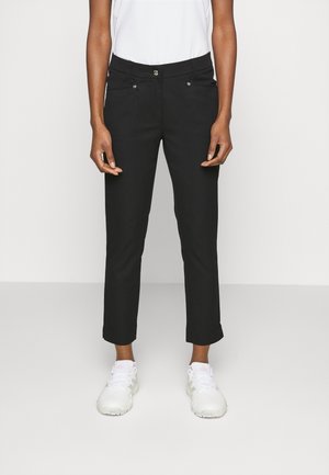 Person, die schwarze, knöchellange Hosen mit Fronttaschen und weiße Turnschuhe trägt, steht vor einem schlichten hellen Hintergrund.