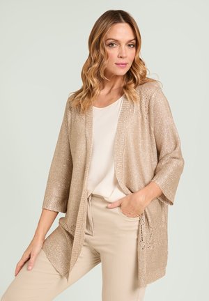Mujer con cárdigan de punto beige sobre top blanco y pantalones beige, posando con una mano en el bolsillo sobre fondo claro.