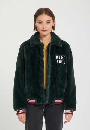 Joven mujer vistiendo una chaqueta peluda verde oscuro con botones en la parte delantera y parche "RIDE FREE" en el pecho, combinada con pantalones oscuros y camiseta amarilla.