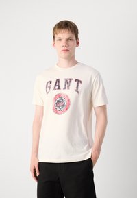 Cream cotton t-shirt featuring a large "GANT" logo in red and blue, with a circular graphic below. Classic crew neck and short sleeves.