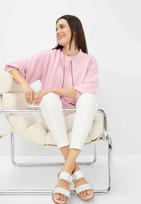 Sweatshirt oversize rose clair avec un détail zippé, associé à un pantalon blanc ajusté et des sandales blanches avec boucle, assis sur un fauteuil moelleux.