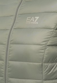 Svijetlozeleni puffer kaput s kvadratnim dijelovima, sa središnjim patentnim zatvaračem i EA7 Emporio Armani logotipom na lijevoj strani prsnog koša.