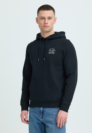 Młody mężczyzna ubrany w czarną bluzę z kapturem z białym napisem na wysokości kieszeni na piersi oraz niebieskie dżinsy, stojący na tle jasnego, jednolitego tła.