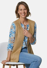 Weste aus hellem Wildleder, ärmelloses Design, über einer blauen Bluse mit floralem Muster getragen. Helle blaue Jeans mit schmaler Passform, auf einem weißen Stuhl sitzend.