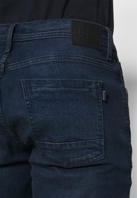 Vaqueros de un azul oscuro con una textura de denim suave, que presentan un bolsillo trasero, costuras sutiles y una etiqueta de marca rectangular de cuero negro.