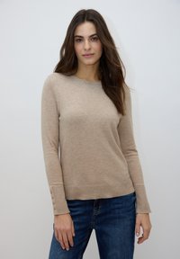 Pull beige à manches longues avec un col rond et un bord-côte. Détails de boutons sur les poignets. Associé à un jean bleu.