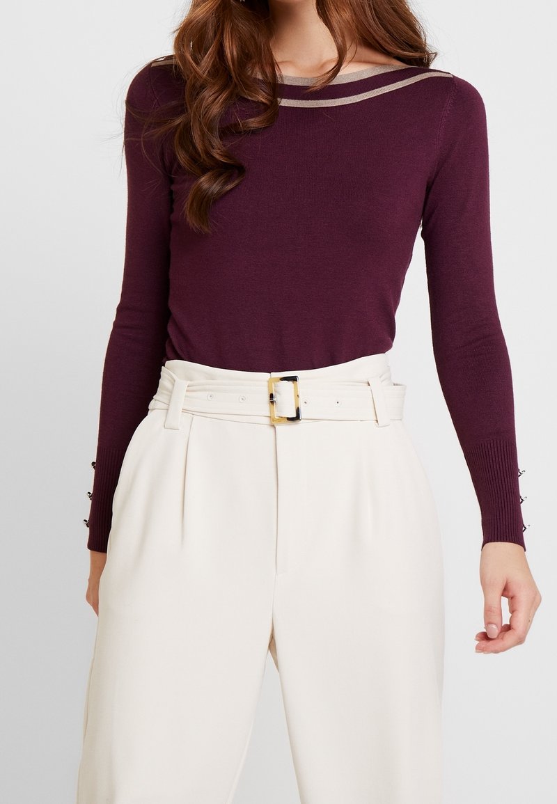 Burgundin pitkähihaisena paita, jossa on beige raidallinen kaula-aukko ja napit korostamassa hihoja. Yhdistetty korkeavyötäröisten kermaisten housujen kanssa.