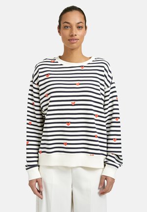 Woman wearing a white and navy striped long-sleeve top with scattered small red heart patterns, paired with white pants.