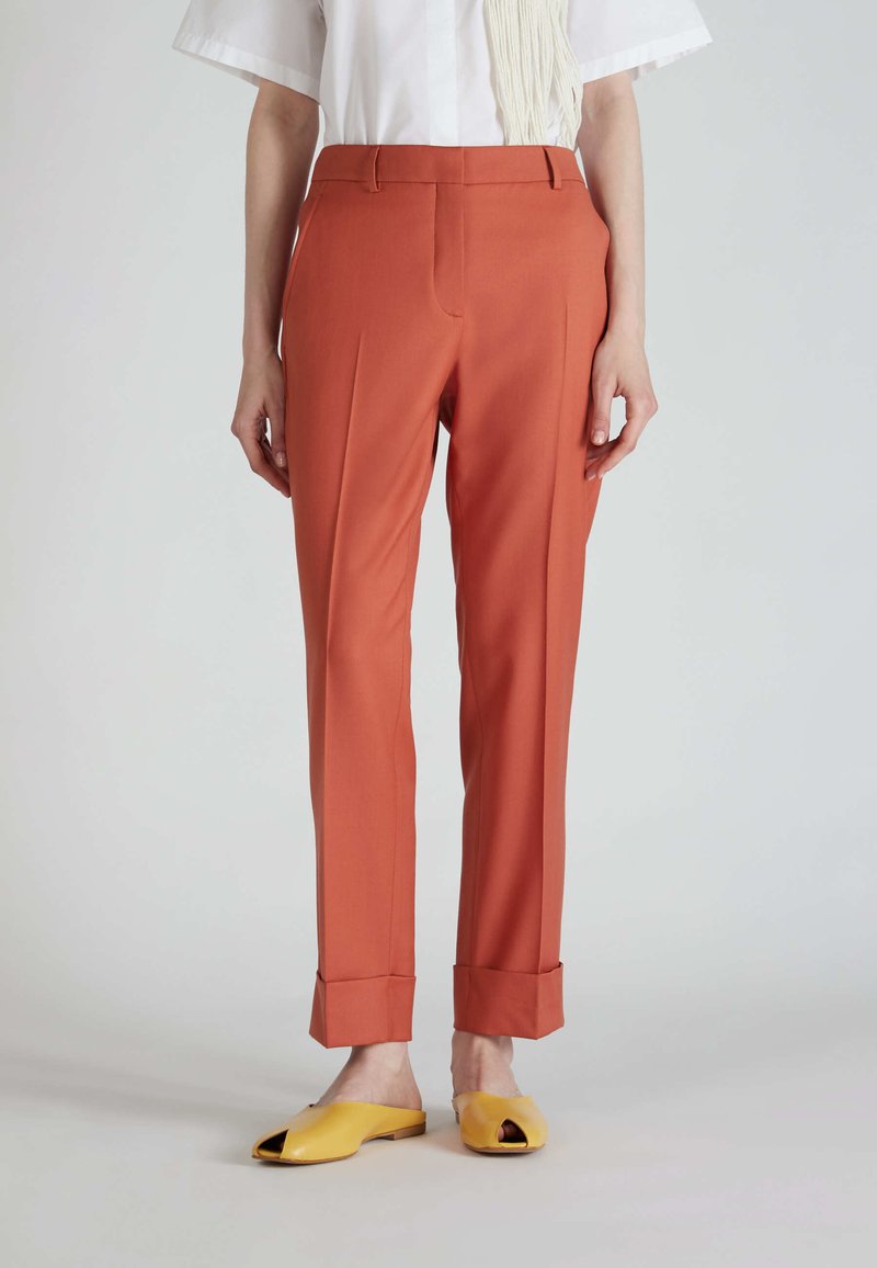 Pantaloni arancioni su misura con gamba dritta, pieghe sottili e risvolti, abbinati a scarpe gialle slip-on. Texture del tessuto liscia.