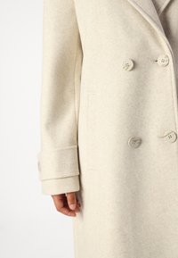 Cappotto in misto lana beige con design doppiopetto, grandi bottoni e una finitura leggermente testurizzata. Include un dettaglio con una tasca singola.