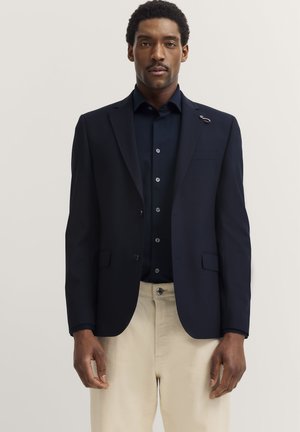 Marineblazer mit Revers und Knopfverschluss, kombiniert mit einem dunklen Hemd. Beigefarbene Hosen. Der Blazer hat eine Nadelakzent am Revers.