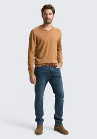 Hellbrauner V-Ausschnitt-Pullover aus weichem Material, kombiniert mit blauen Jeans und braunen Wildleder-Stiefeletten. Schlichtes, tailliertes Design mit klaren Linien.