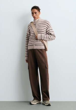 Une femme porte une veste en maille rayée, un pantalon cargo marron, des baskets et un sac beige en bandoulière, devant un mur de studio uni.