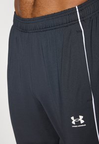 Melni sporta šorti ar elastīgu jostasvietu, ar baltu sānu svītru un balto Under Armour logotipu apakšējā kājā.
