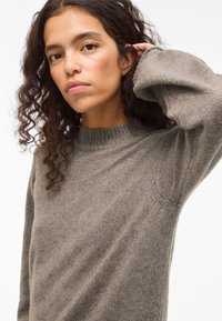 Ženska s kodrastimi temnimi lasmi, oblečena v ohlapen siv pleteni pulover, dviguje eno roko, da se dotakne svojih las, pred enostavno belo ozadjem.