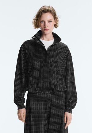 Młoda osoba z krótkimi włosami nosi czarną kurtkę zapinaną na suwak z wzorem w kratę oraz dopasowane spodnie, stoi na tle gładkiego, jasnego tła.