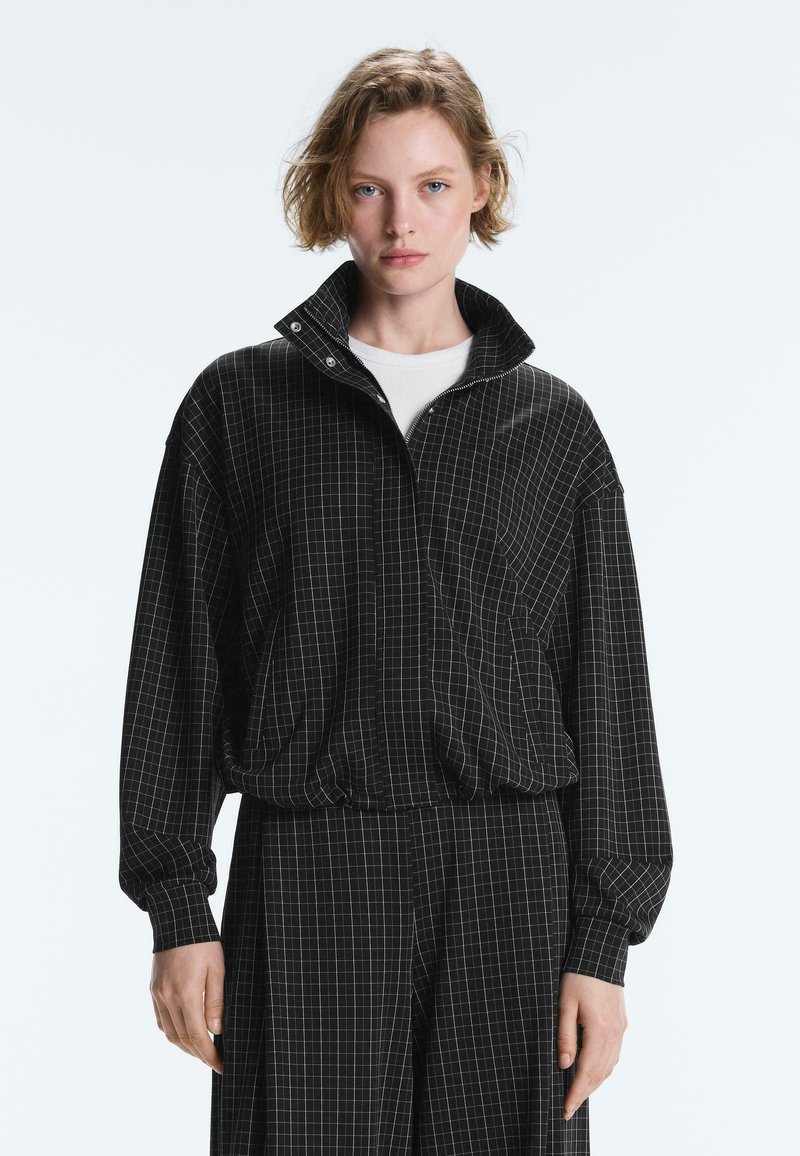 Jeune personne aux cheveux courts porte une veste noire zippée à motif quadrillé et un pantalon assorti, debout devant un fond clair uni.