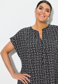 Tunic nera con motivo a zigzag bianco, maniche corte e scollatura a V. Tessuto morbido con vestibilità rilassata e dettagli minimi.