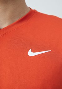 Červené sportovní tričko vyrobené z texturované tkaniny s designem ve tvaru V a bílým logem Nike na levé části hrudi.