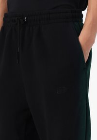 Černé tepláky s šňůrkou v pase, s bočními kapsami a jemným logem, vyrobené z měkké látky s hladkým povrchem.