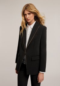 Blazer negro con un diseño estructurado, que presenta detalles de cremallera dorada, un bolsillo en el pecho y dos bolsillos laterales. Estilizado con una camisa blanca debajo.