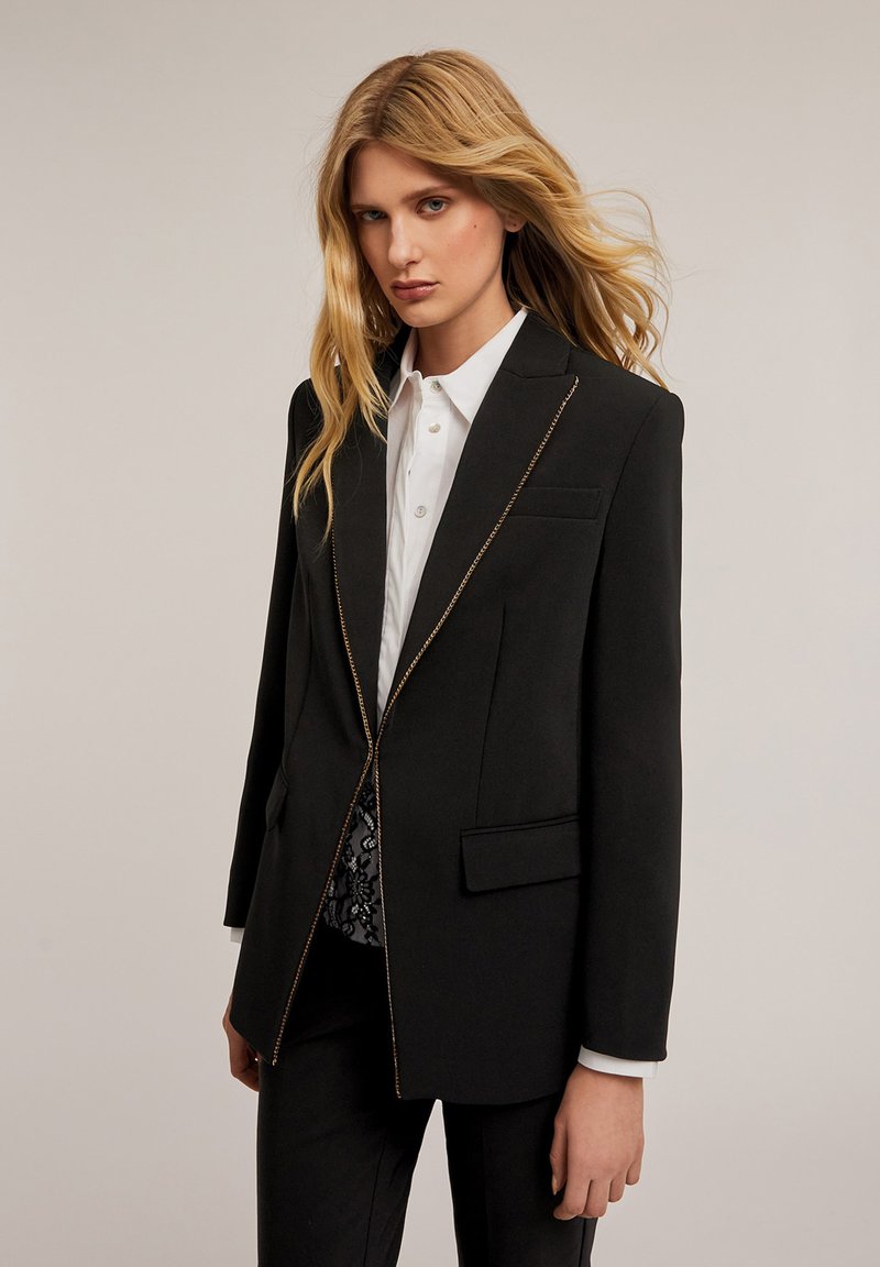 Blazer negro con un diseño estructurado, que presenta detalles de cremallera dorada, un bolsillo en el pecho y dos bolsillos laterales. Estilizado con una camisa blanca debajo.
