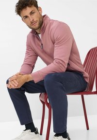 Pullover a mezza zip in tessuto rosa chiaro, abbinato a pantaloni blu scuro. L'uomo è seduto su una sedia rossa, indossando sneakers bianche e nere.