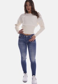 Maglione lavorato a maglia color crema con orlo a coste, abbinato a jeans blu aderenti. Il modello indossa sneaker bianche con dettagli neri.