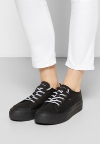 Svarta canvas-sneakers med rund tå, vita skosnören och en strukturerad svart gummisula. Signaturlogotyp på tungan. Vita byxor i ankle-längd.