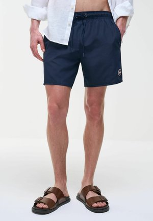 Uomo che indossa pantaloncini da bagno blu navy con coulisse, camicia bianca sbottonata e sandali marroni con fibbia, in piedi su sfondo bianco.