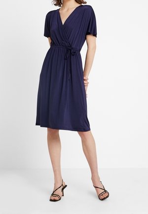 Femme portant une robe portefeuille bleu marine mi-longue avec manches courtes et taille nouée, associée à des sandales à talons noires avec brides.