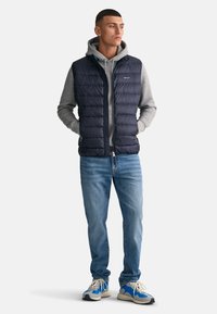 Chaleco de plumas azul marino con diseño acolchado sobre una sudadera gris, combinado con jeans azul claro y zapatillas con acentos azules.